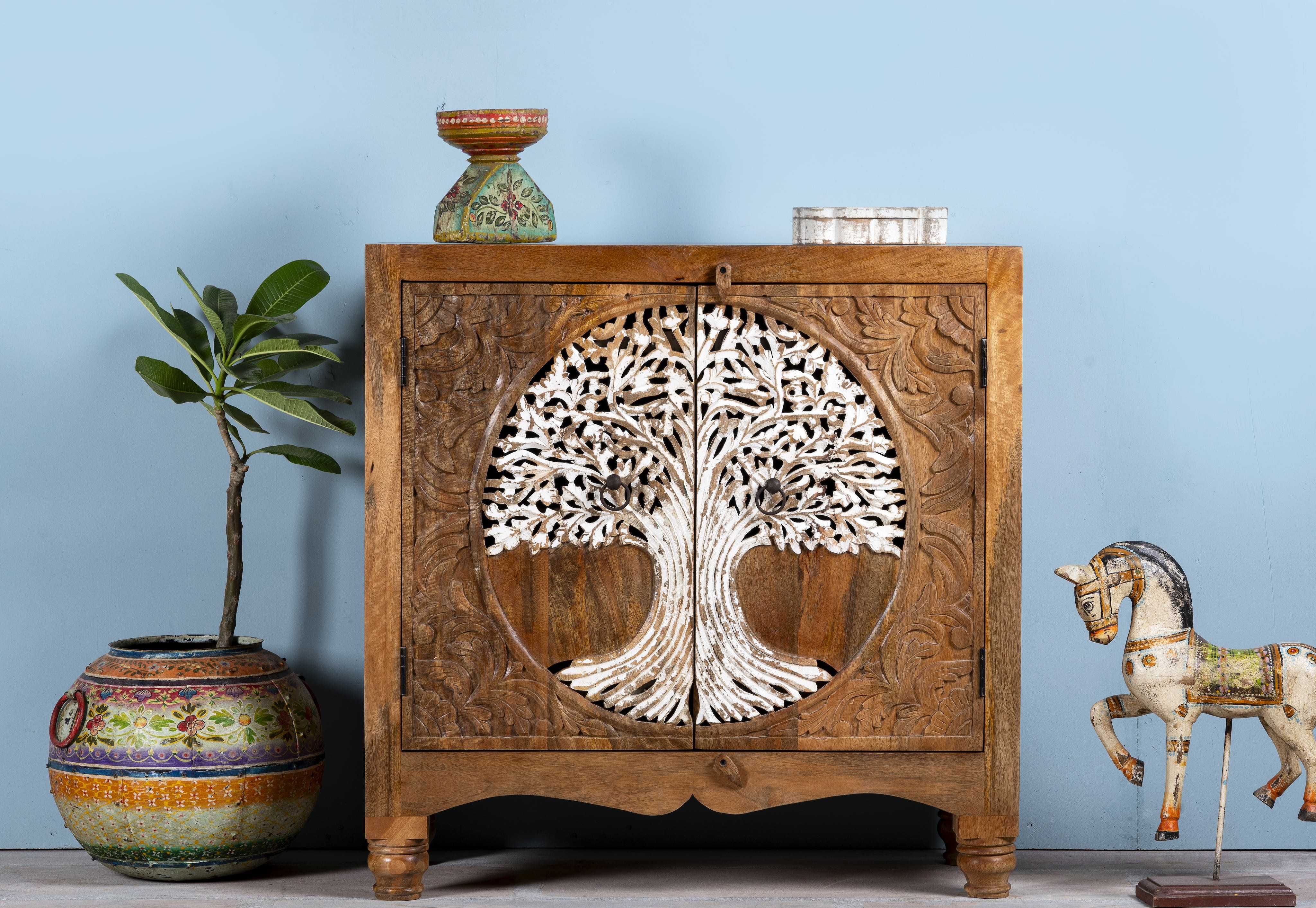 Solid mango wood cupboard with hand-carved Tree of Life design and whitewashed finish, featuring two doors and a curved base, placed against a blue wall with decorative items around it.