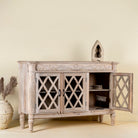 Side angle image with open door of a rustic sideboard made of Mango wood with hand-carved details and glass panelled doors featuring a diamond lattice design.