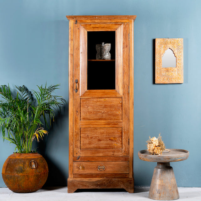 An antique Indian teak wood showcase with a glass door and bronze-colored hardware, featuring interior shelves and a drawer, placed against a blue wall next to a potted plant and a decorative mirror.