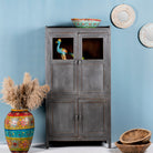 A refurbished teak wood showcase cabinet with glass doors and a shabby chic grey finish, displayed in a home setting with decorative items inside and around it.