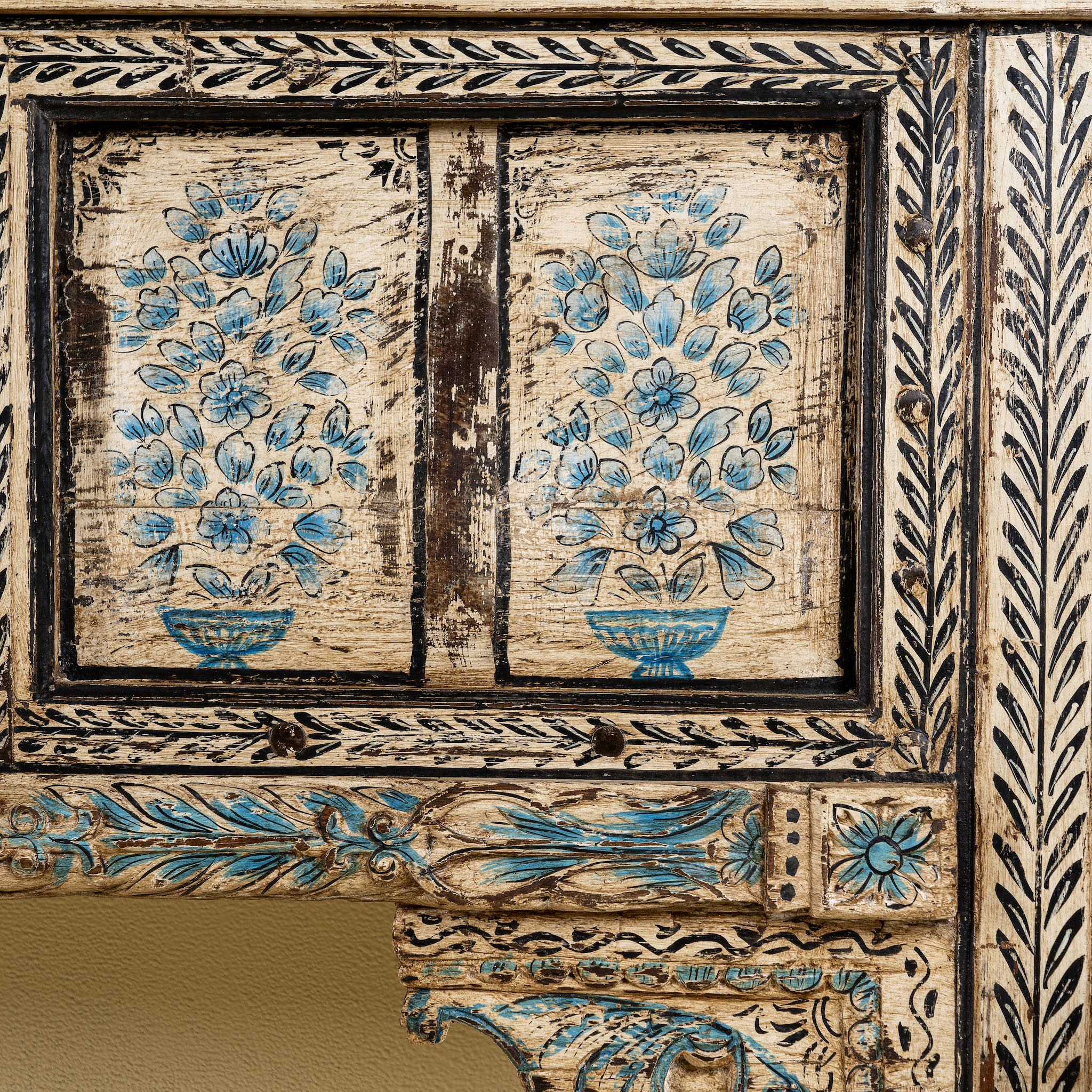 Close up image of a  hand-painted console table made from reclaimed teak wood, featuring intricate floral motifs in blue tones across the front panels, framed by carved detailing and a distressed finish that highlights its artisanal and vintage character.
