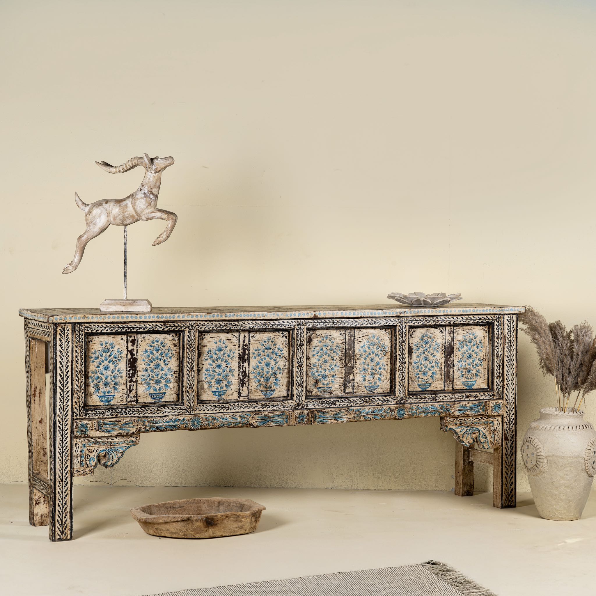 Side angle image of a hand-painted console table made from reclaimed teak wood, featuring intricate floral motifs in blue tones across the front panels, framed by carved detailing and a distressed finish that highlights its artisanal and vintage character.
