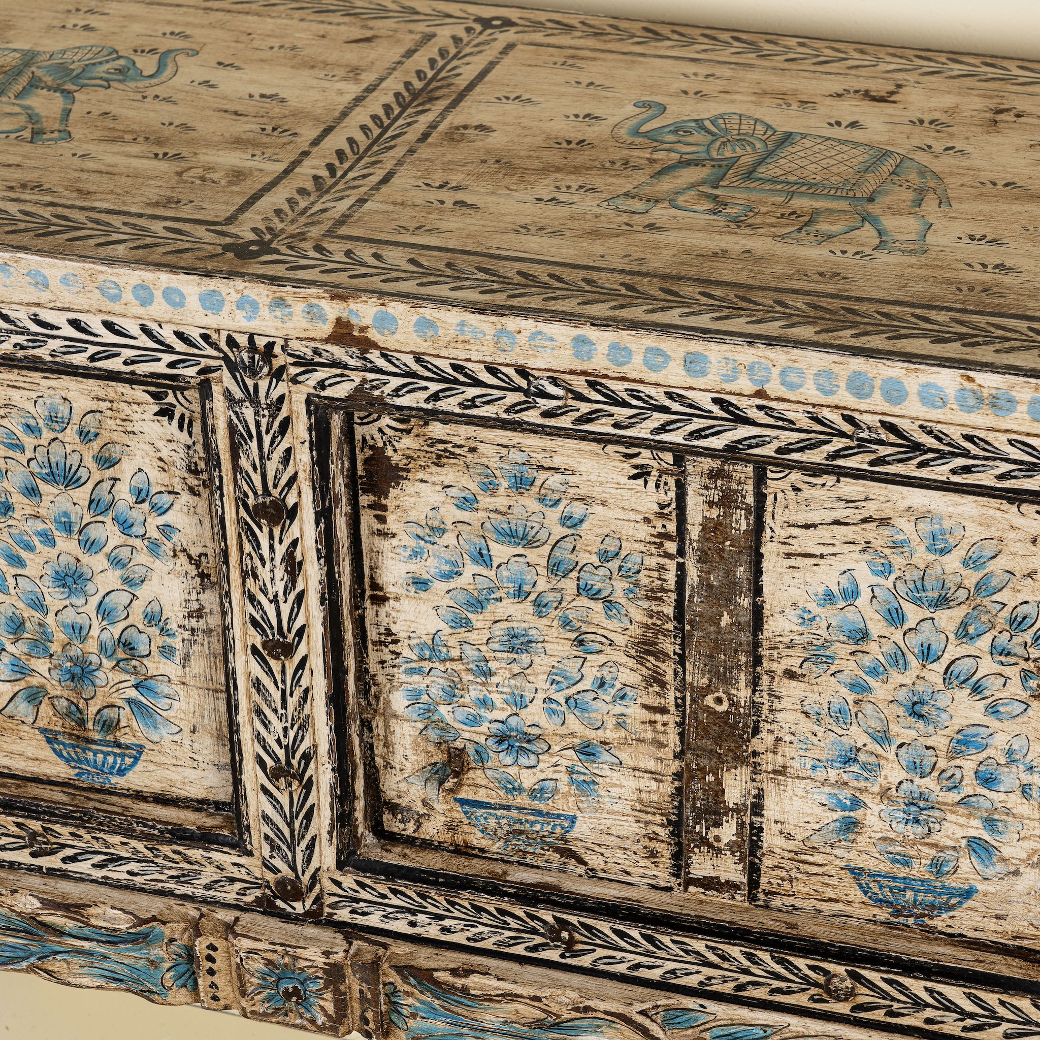 Close up side image of a hand-painted console table made from reclaimed teak wood, featuring intricate floral motifs in blue tones across the front panels, framed by carved detailing and a distressed finish that highlights its artisanal and vintage character.
