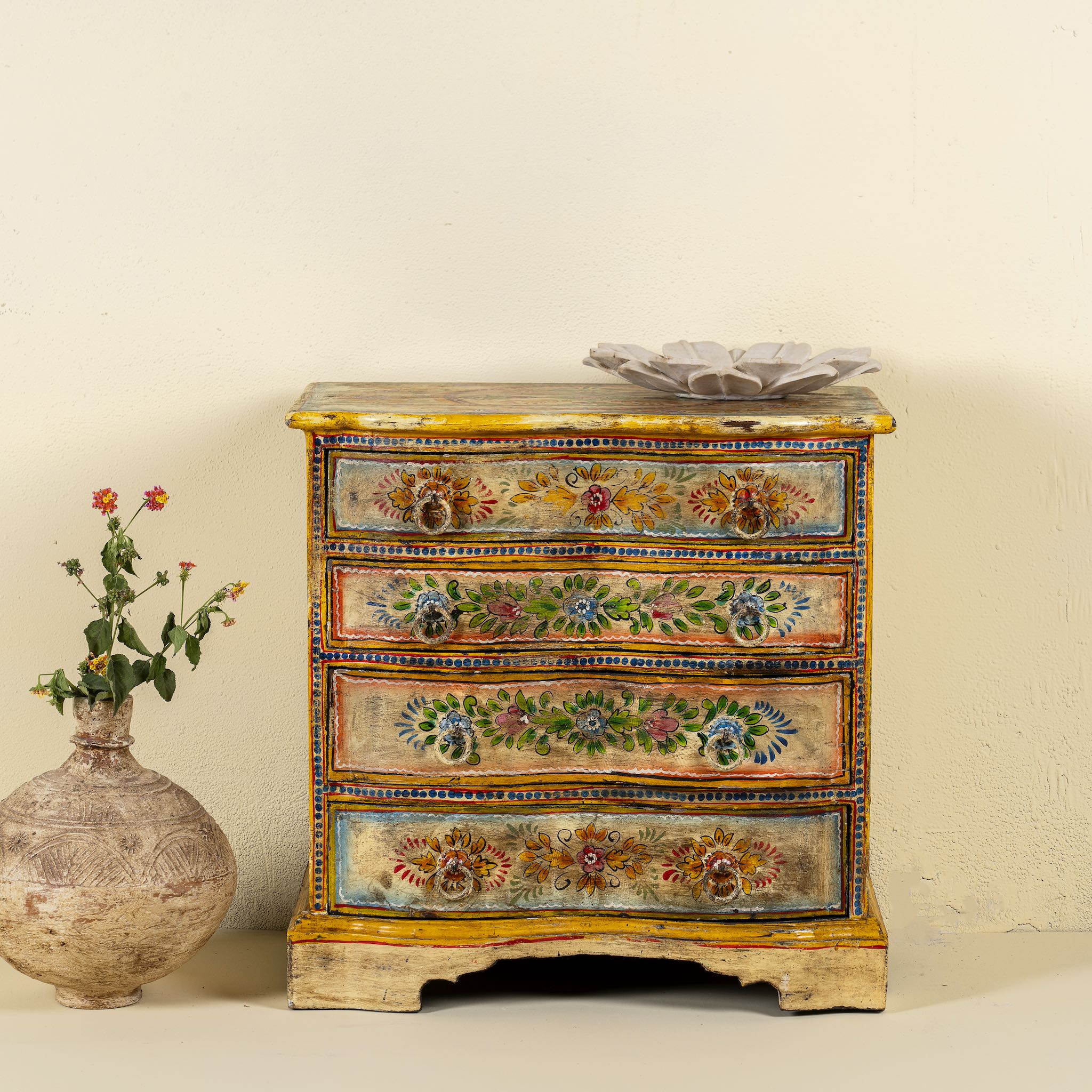 Front view image of a hand-painted chest of drawers made from reclaimed wood, featuring a vibrant floral motif across its four drawers, with a distressed finish and colourful detailing that highlights its vintage, artisanal charm.
