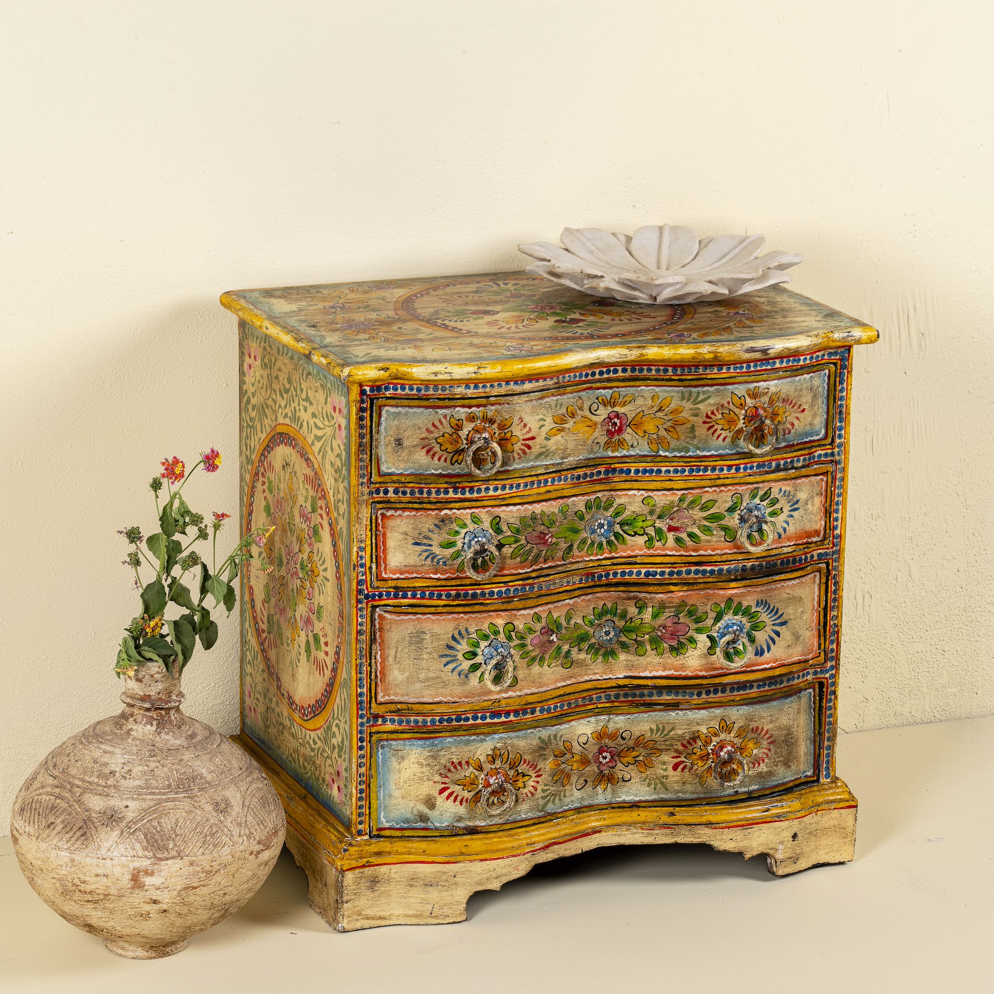 Side angle image of a hand-painted chest of drawers made from reclaimed wood, featuring a vibrant floral motif across its four drawers, with a distressed finish and colourful detailing that highlights its vintage, artisanal charm.
