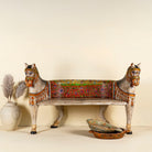Front view image of a hand-painted bench made from reclaimed wood, designed with intricately carved double horse heads at each end, featuring a vibrant floral backrest and detailed decorative patterns, blending traditional craftsmanship with whimsical artistry.
