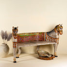Side angle image of a hand-painted bench made from reclaimed wood, designed with intricately carved double horse heads at each end, featuring a vibrant floral backrest and detailed decorative patterns, blending traditional craftsmanship with whimsical artistry.
