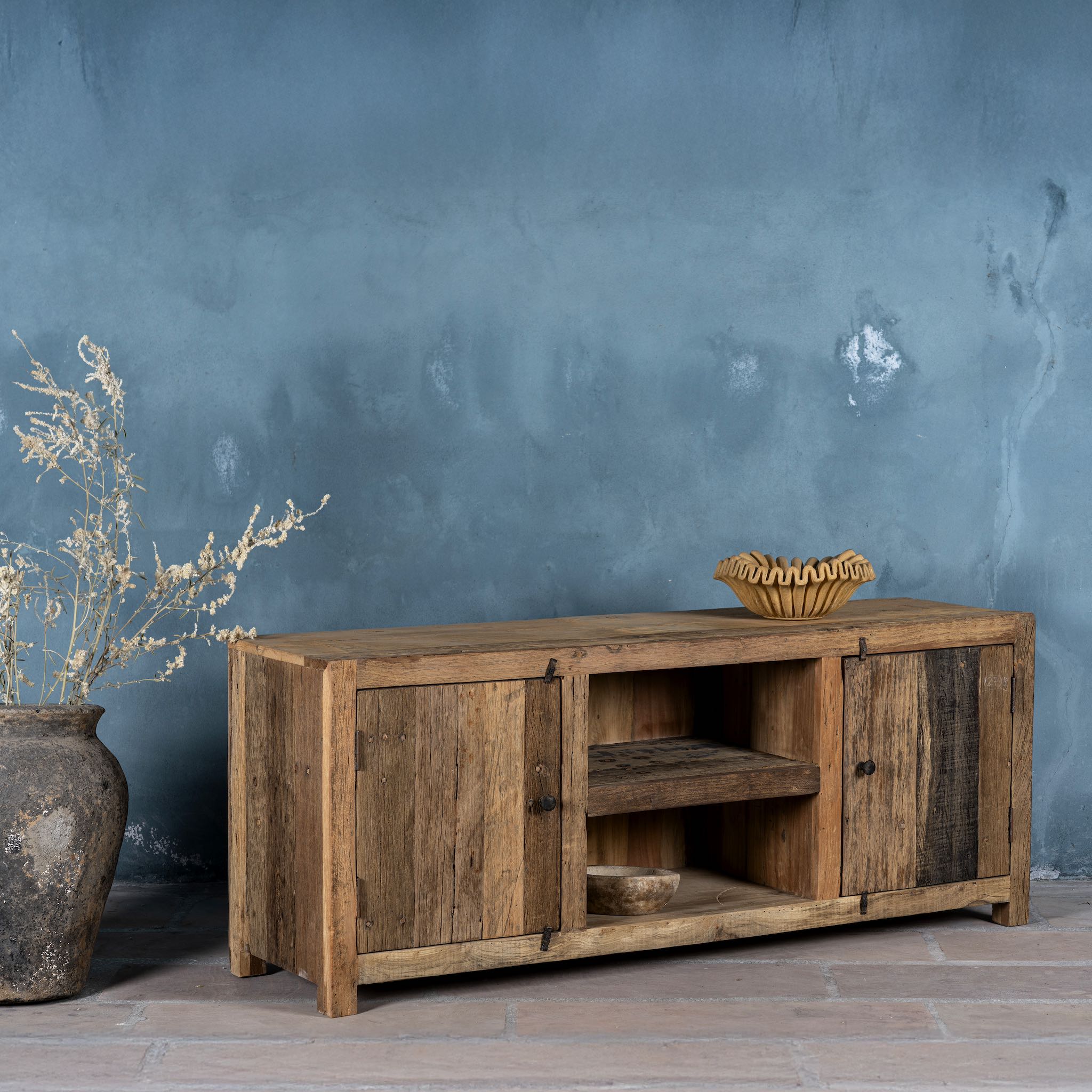 Side view image of a rustic sideboard in Driftwood/Wabi Sabi style made from recycled old wood, featuring a natural, weathered finish with two cabinet doors and open central shelving.


