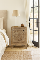 Lifestyle scene of a pair of hand-carved bedside tables made of mango wood, featuring intricate round floral motifs on the doors, crackled vintage finish, and elegant curved legs in a distressed antique design.