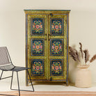 A vibrantly hand-painted reclaimed wood vintage cabinet with floral motifs, in a colorful and traditional design, standing in a room with a chair and a decorative vase beside it.