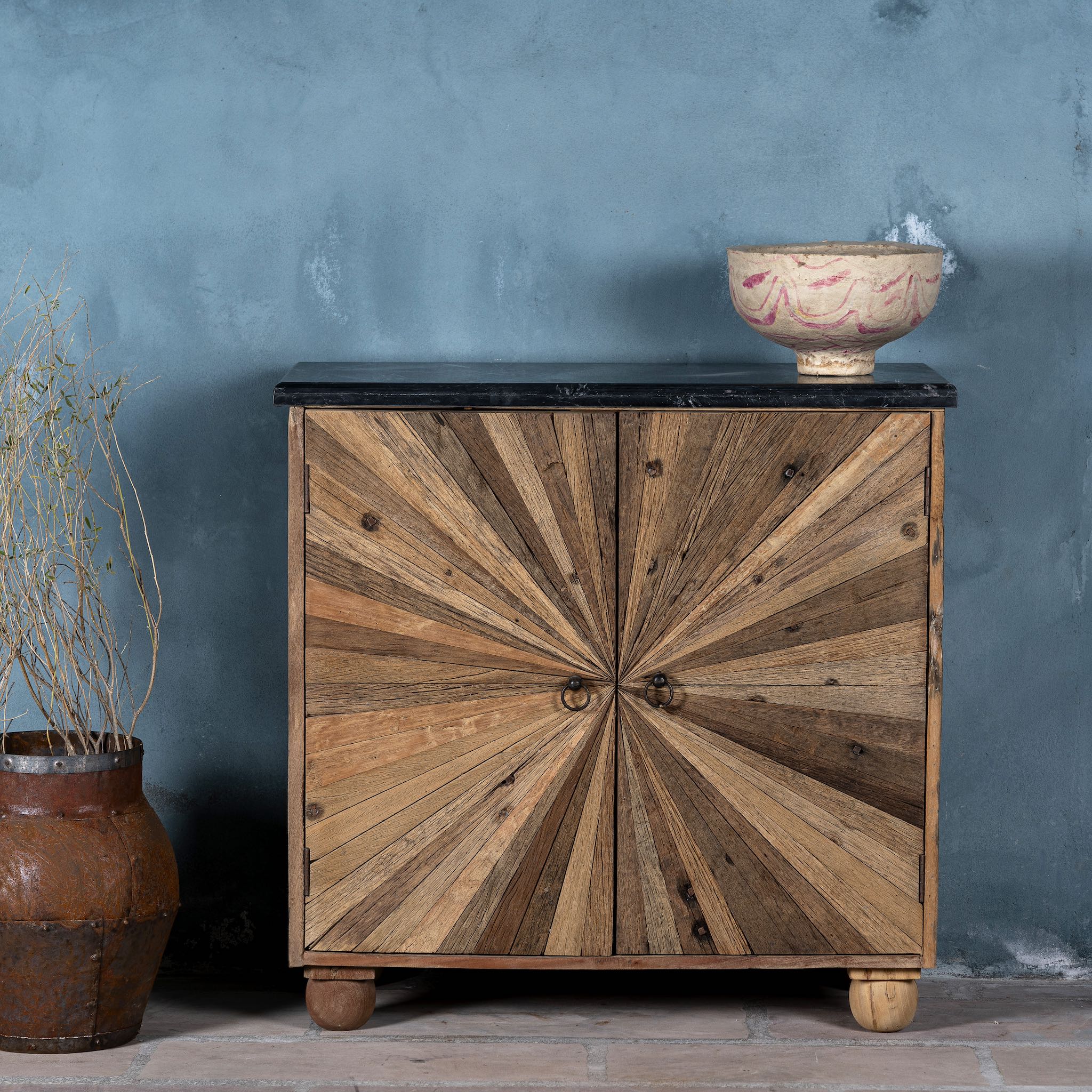 front view image of a rustic-style cabinet made from reclaimed wood, featuring a striking sunburst pattern on the double doors, round metal pulls, and a contrasting black marble top that adds a refined touch to its natural, weathered look.
