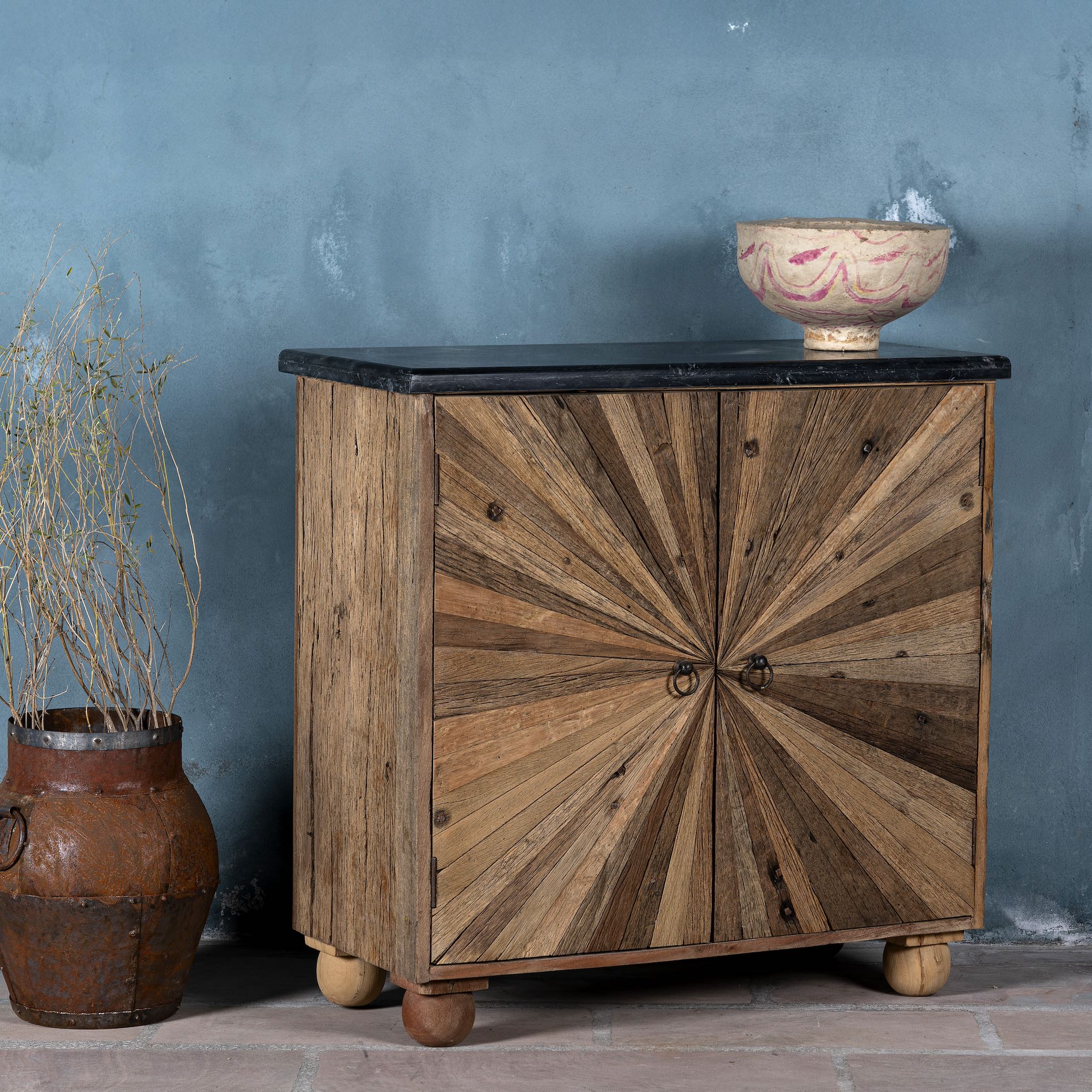 Side angle image of a rustic-style cabinet made from reclaimed wood, featuring a striking sunburst pattern on the double doors, round metal pulls, and a contrasting black marble top that adds a refined touch to its natural, weathered look.
