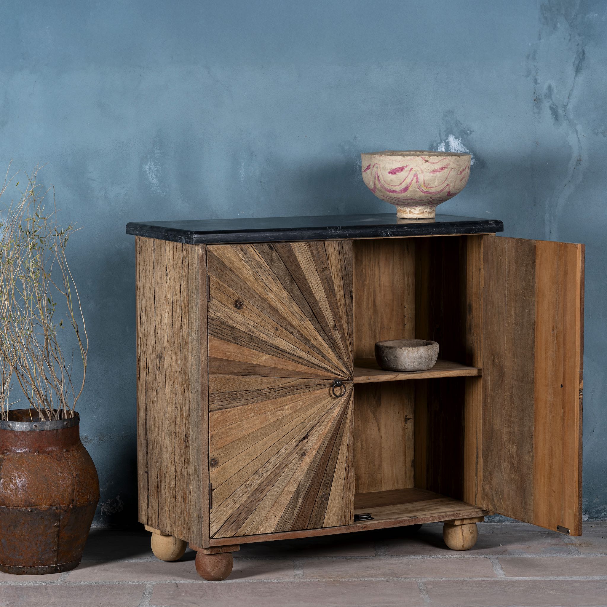 Open door image of a  rustic-style cabinet made from reclaimed wood, featuring a striking sunburst pattern on the double doors, round metal pulls, and a contrasting black marble top that adds a refined touch to its natural, weathered look.
