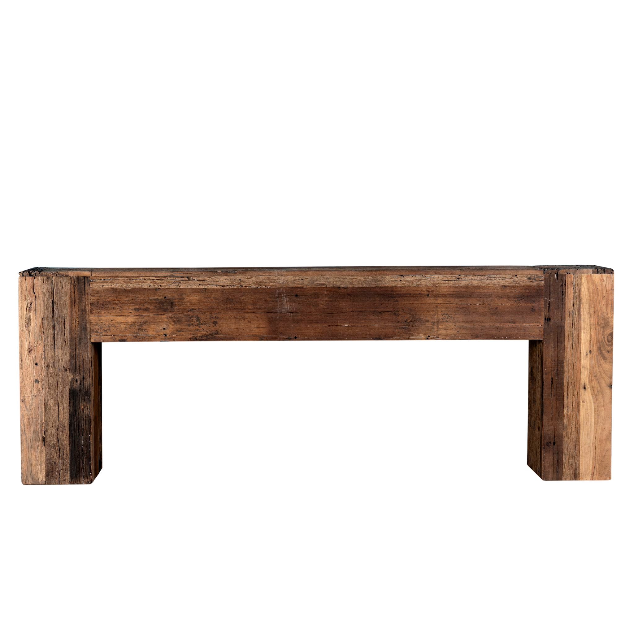 White background image of a large rustic console table made from reclaimed wood, showcasing a solid rectangular design with thick block legs and a rich, weathered finish; styled against a textured blue wall with decorative accents.
