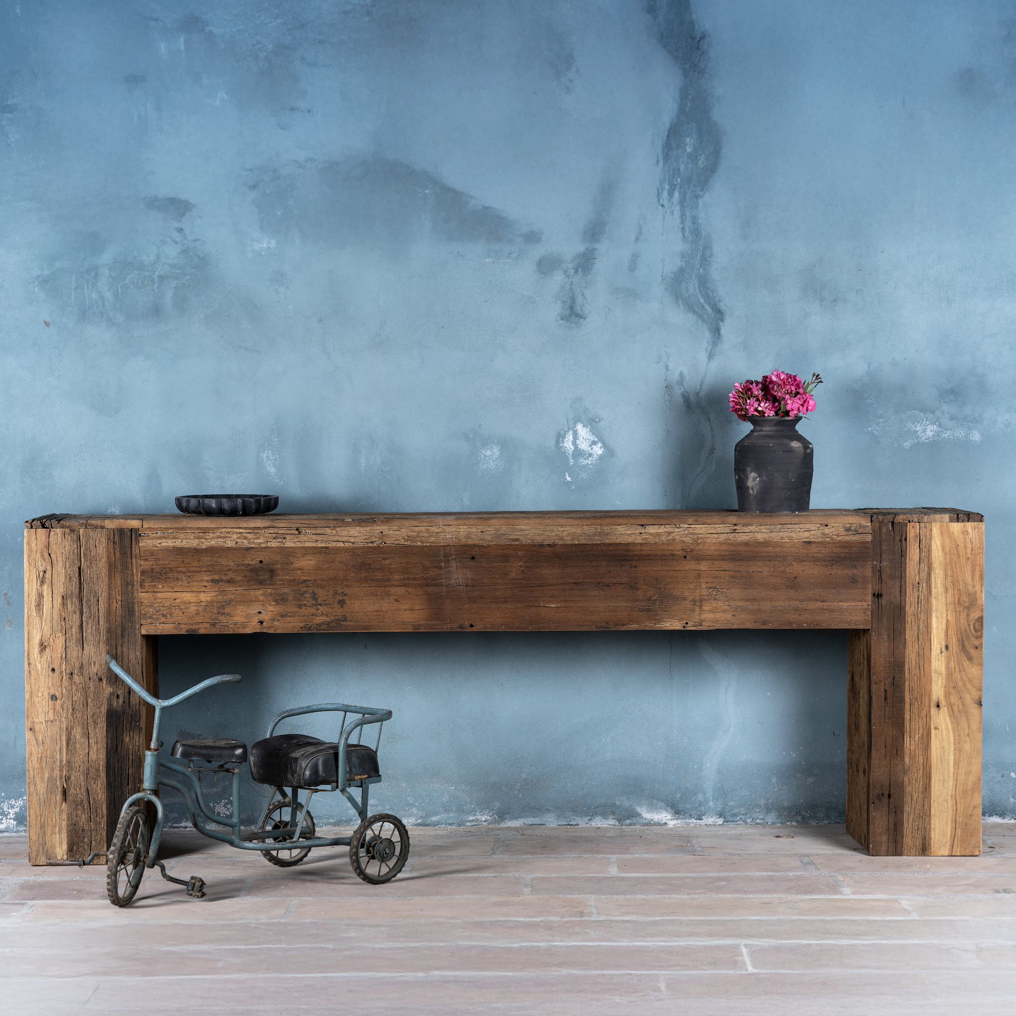 Front view image of a large rustic console table made from reclaimed wood, showcasing a solid rectangular design with thick block legs and a rich, weathered finish; styled against a textured blue wall with decorative accents.
