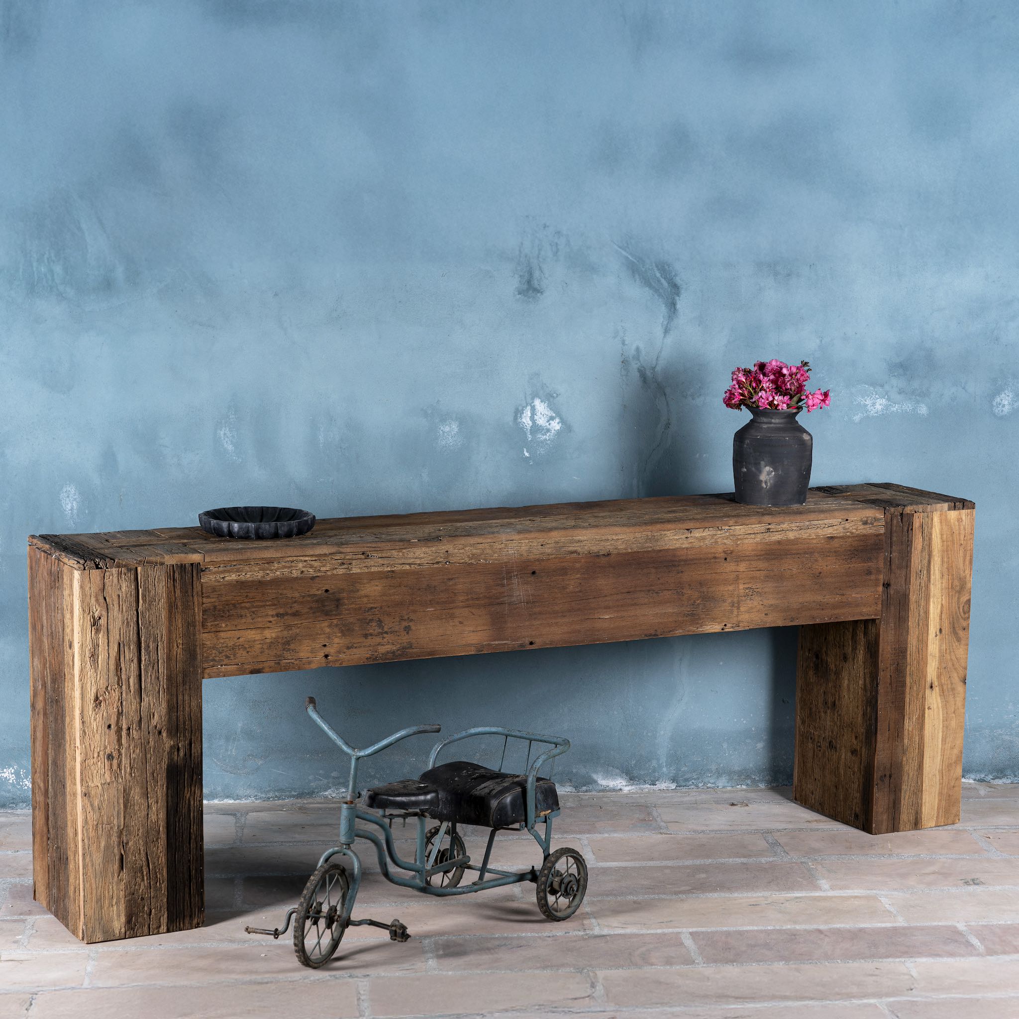 Side image of a  large rustic console table made from reclaimed wood, showcasing a solid rectangular design with thick block legs and a rich, weathered finish; styled against a textured blue wall with decorative accents.
