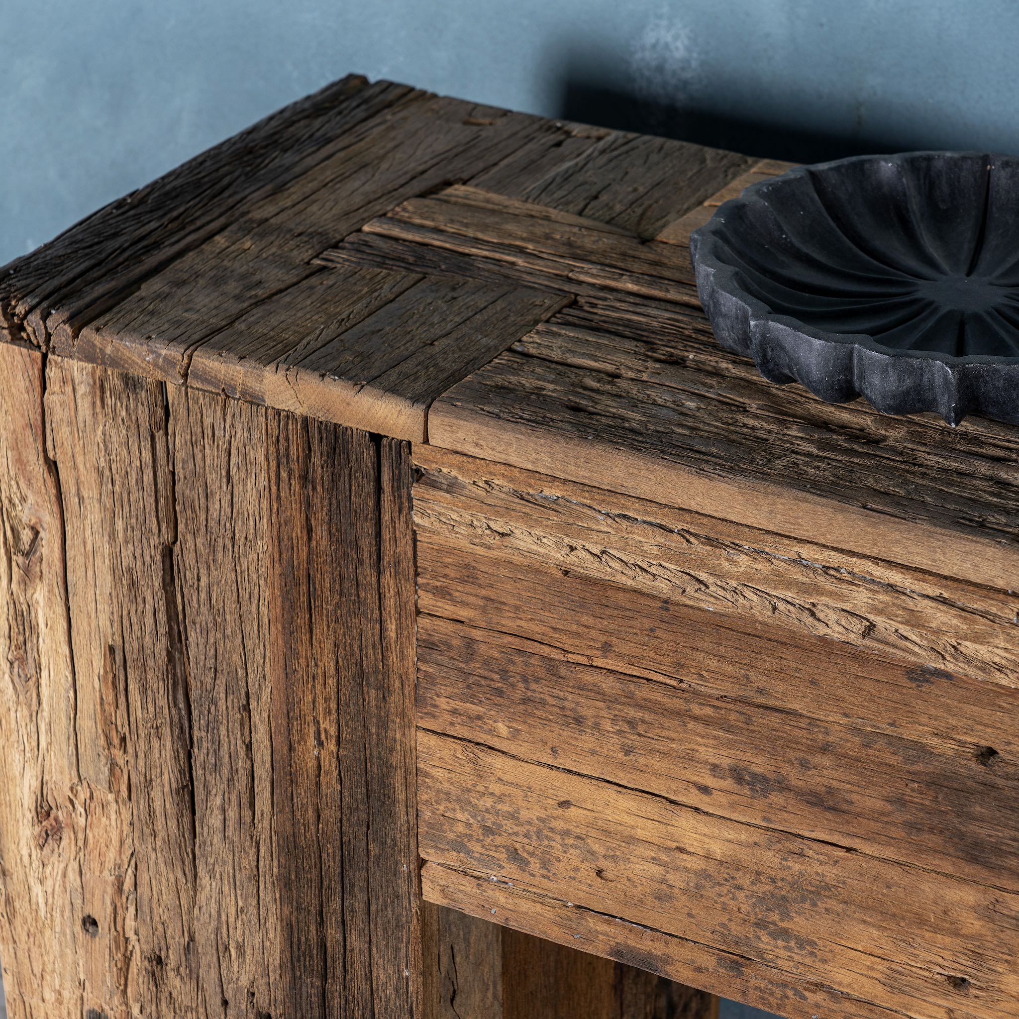 Close up image of a large rustic console table made from reclaimed wood, showcasing a solid rectangular design with thick block legs and a rich, weathered finish; styled against a textured blue wall with decorative accents.
