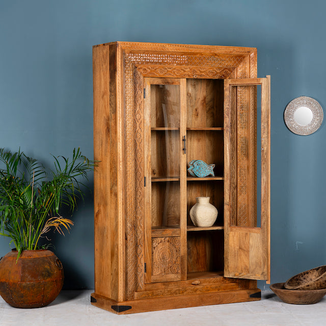 Side angle image with open doors of a hand carved display cabinet made from natural mango wood with floral detailing and glass-panelled doors; 180 × 110 × 40 cm.