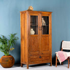 Side angle image of a tall reclaimed teak wood display cabinet with two glass doors and a bottom drawer – wooden display cabinet crafted as sustainable furniture with natural elegance.