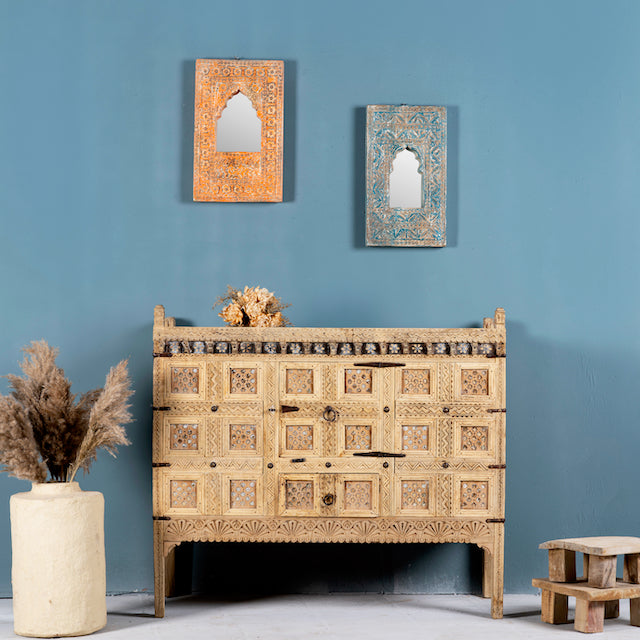 An antique Indian treasure chest with sandblasted surface and hand-carved patterns, featuring iron applications, displayed against a blue wall with decorative items and a vase beside it.