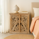 Wooden nightstand with intricate carvings next to a bed in a bedroom.
