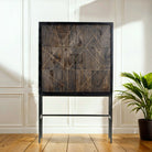 An elegant wooden cabinet with two doors and geometric handcrafted copper inlays, paired with a gray/black nickel-plated metal stand, positioned in a room with a plant to the side.