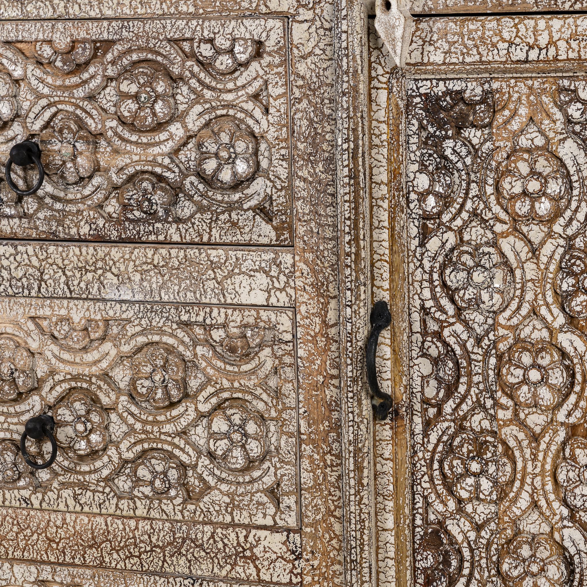 Close up image of a hand-carved sideboard made from mango wood, featuring intricate geometric and floral patterns across the drawers and cabinet door, with a distressed whitewashed finish that enhances its vintage artisanal appeal.
