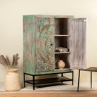 Open door image of a carved mango wood cabinet with floral hand-carving, distressed green finish, iron stand, 100 × 40 × 150 cm.
