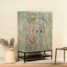 Side angle image of a carved mango wood cabinet with floral hand-carving, distressed green finish, iron stand, 100 × 40 × 150 cm.