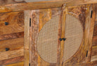 Close up image of a hand-carved mango wood sideboard with rattan inserts, featuring two cabinet doors with rattan fronts and six drawers, placed against a neutral backdrop with decorative items on top.