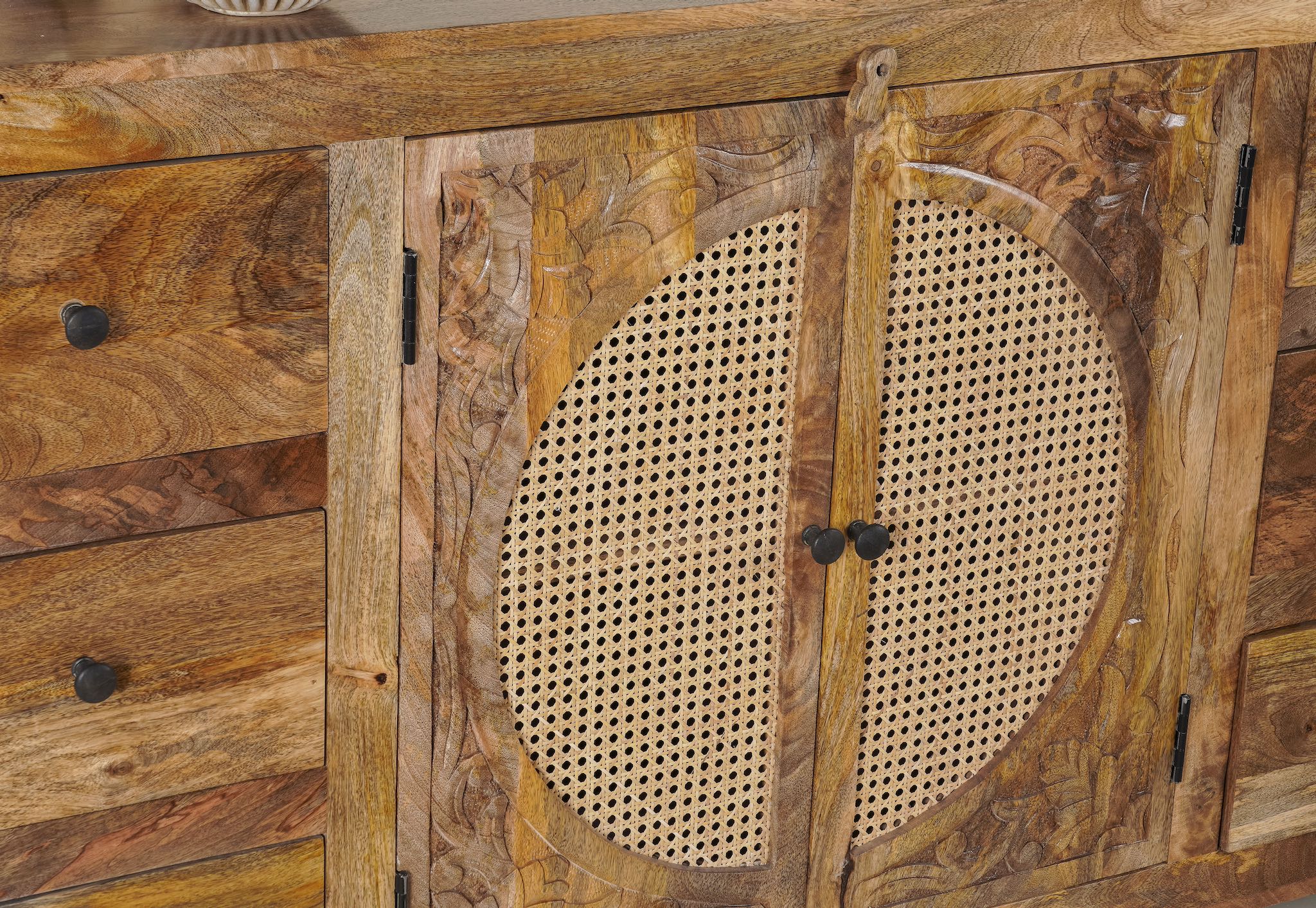 Close up image of a hand-carved mango wood sideboard with rattan inserts, featuring two cabinet doors with rattan fronts and six drawers, placed against a neutral backdrop with decorative items on top.