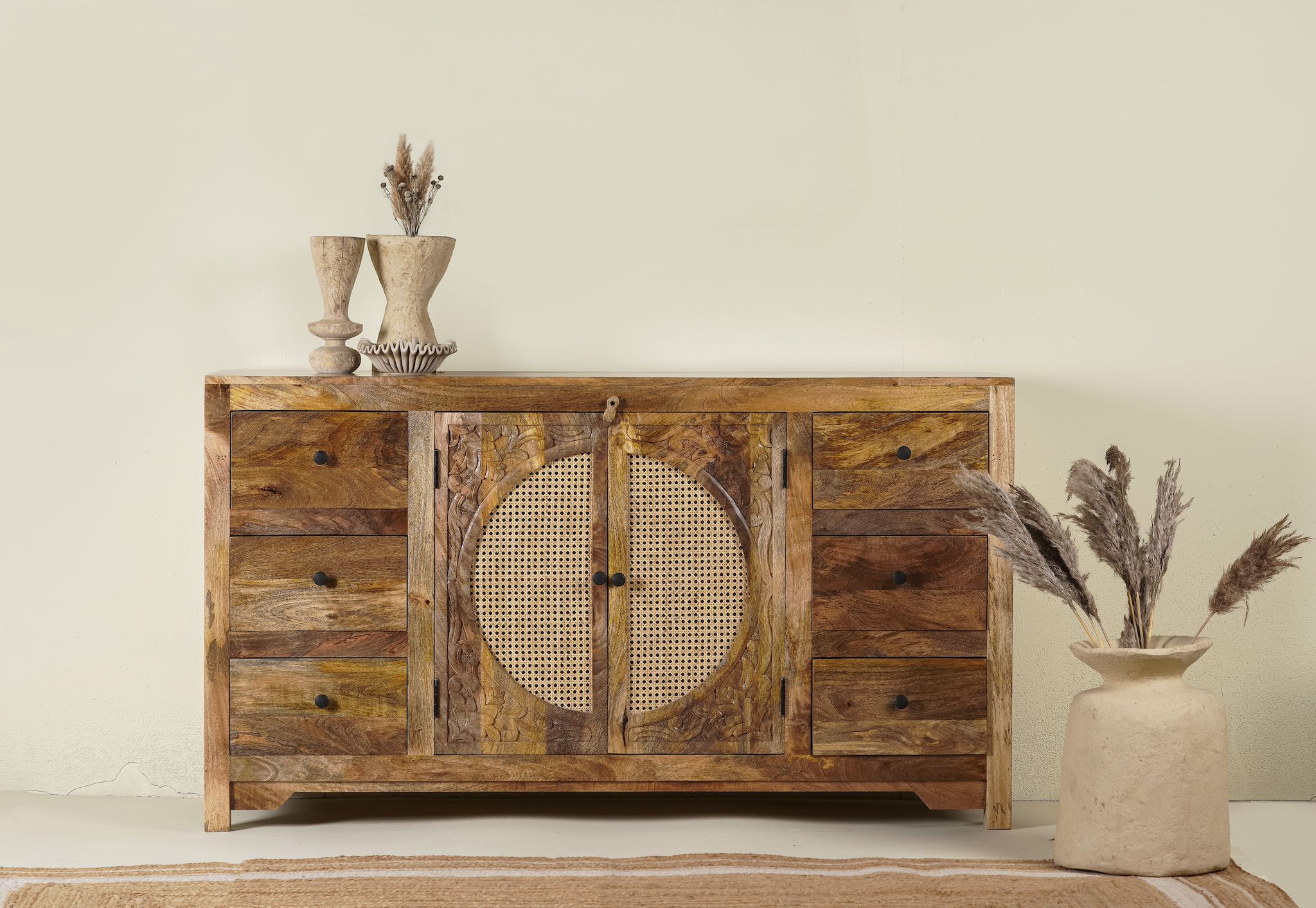 Hand-carved mango wood sideboard with rattan inserts, featuring two cabinet doors with rattan fronts and six drawers, placed against a neutral backdrop with decorative items on top.