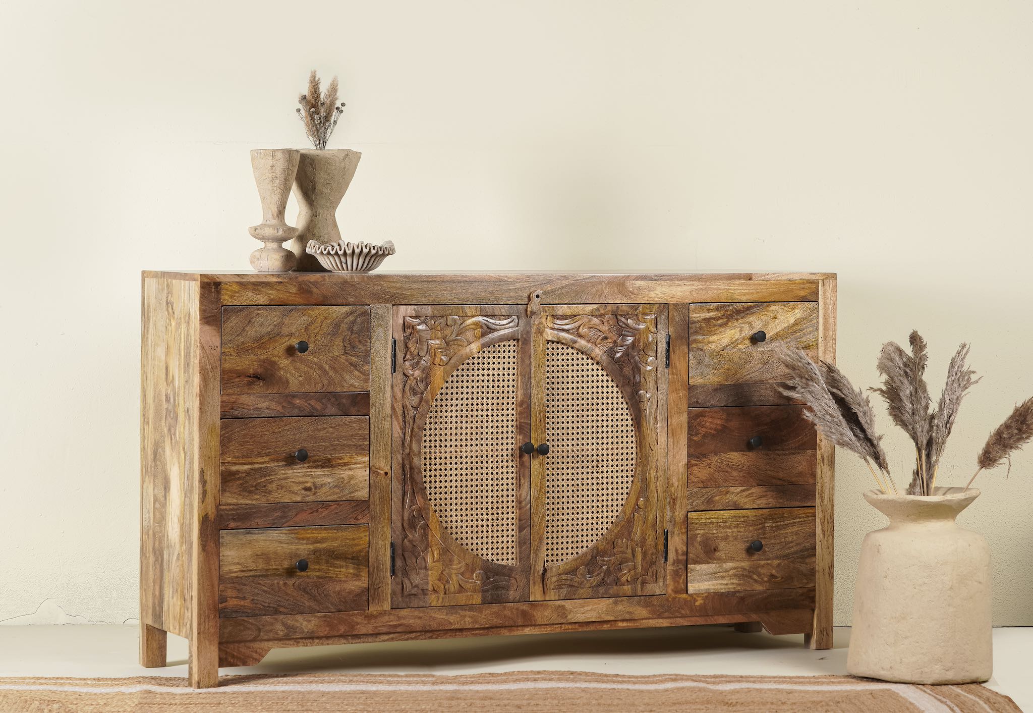 Side angle image of a hand-carved mango wood sideboard with rattan inserts, featuring two cabinet doors with rattan fronts and six drawers, placed against a neutral backdrop with decorative items on top.