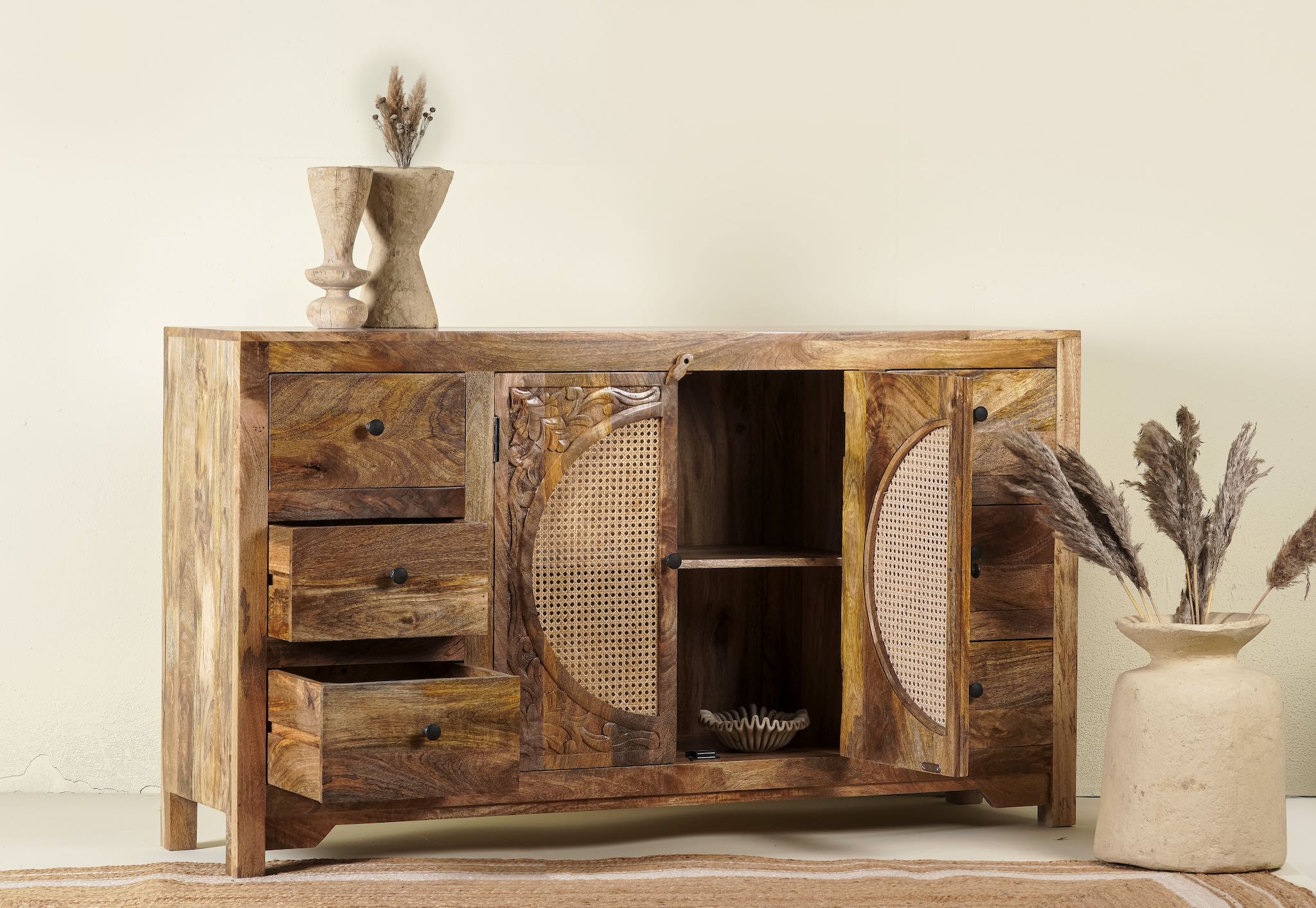 Open door image of a hand-carved mango wood sideboard with rattan inserts, featuring two cabinet doors with rattan fronts and six drawers, placed against a neutral backdrop with decorative items on top.