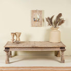 Rustic coffee table Ritvik made from old reclaimed teak wood, featuring a weathered finish, turned legs, decorative carved apron, and visible metal rivets, styled with earthy decor in a minimal interior setting.
