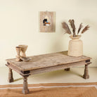 Side angle of rustic coffee table Ritvik made from old reclaimed teak wood, featuring a weathered finish, turned legs, decorative carved apron, and visible metal rivets, styled with earthy decor in a minimal interior setting.
