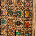Close up image of a tall vintage wall panel made from reclaimed teak wood, intricately hand-painted with a grid of vibrant deity faces, animals, and symbolic motifs in rich colors, each framed within carved squares, reflecting traditional Indian folk art.
