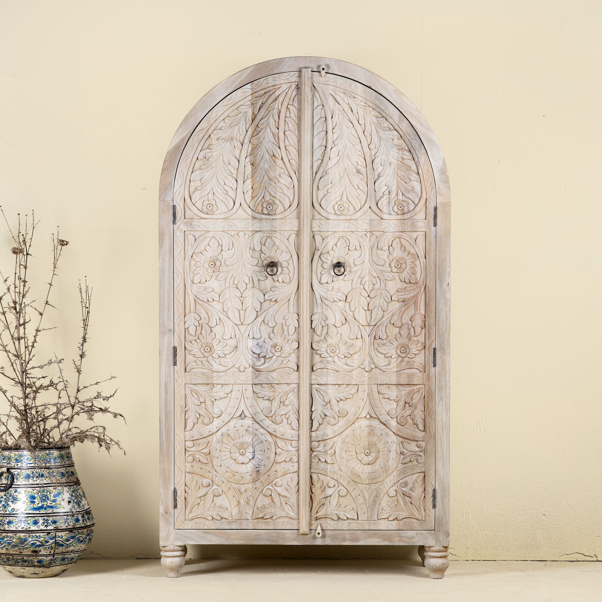 Arched wardrobe made from solid mango wood, featuring intricate hand-carved floral and leaf motifs on double doors, finished in a soft natural tone- ideal for Ibiza or boho interiors.