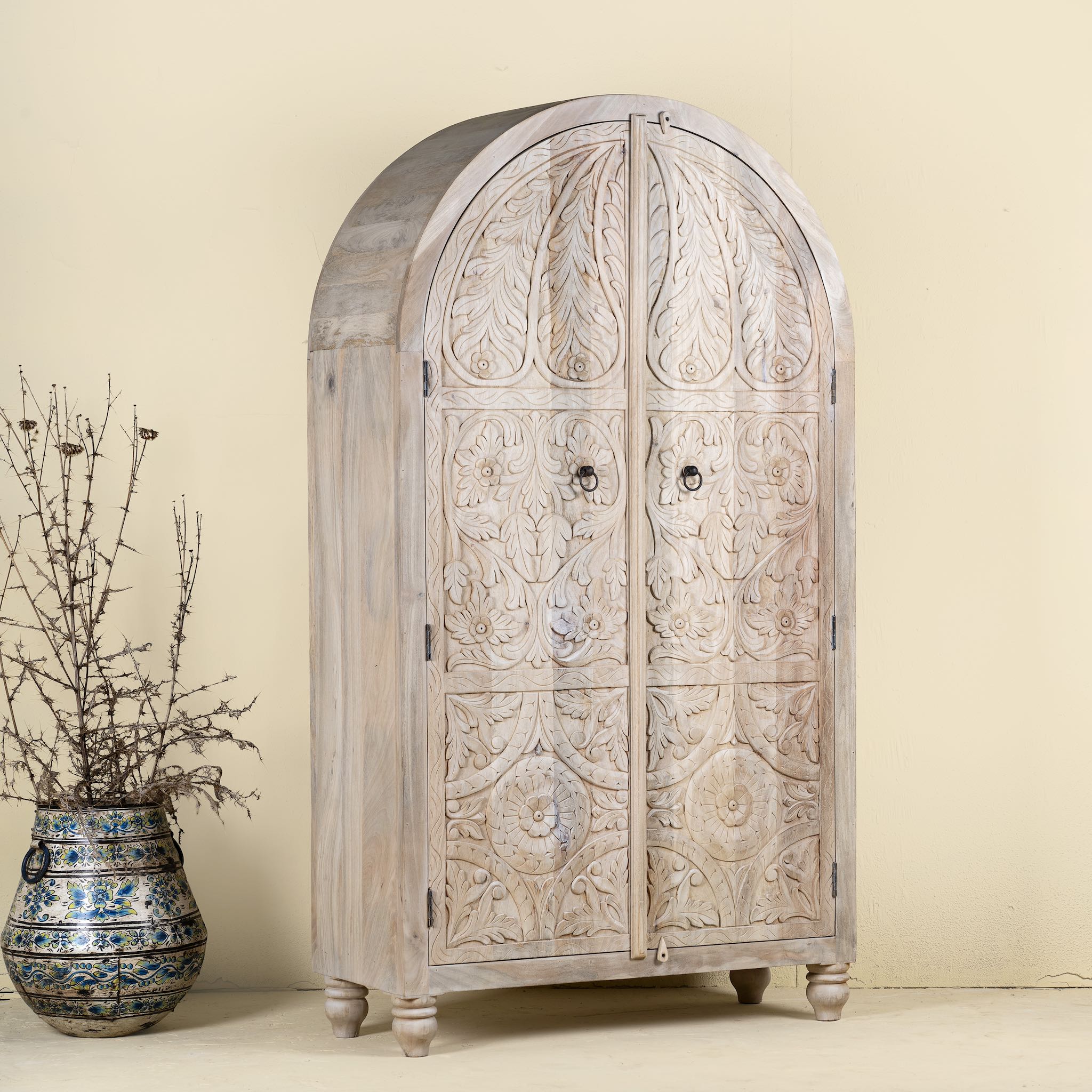 Side angle view of a arched wardrobe made from solid mango wood, featuring intricate hand-carved floral and leaf motifs on double doors, finished in a soft natural tone- ideal for Ibiza or boho interiors.