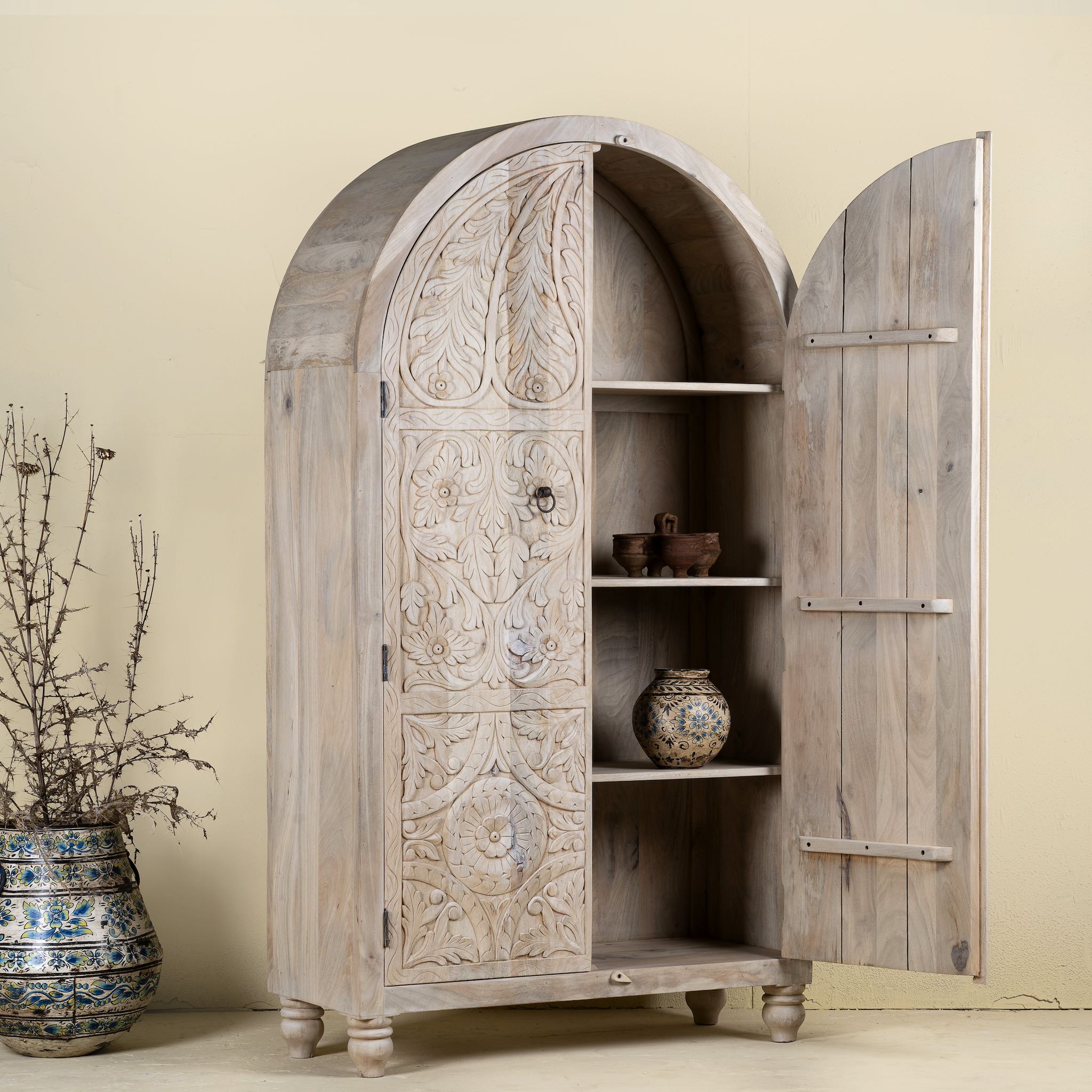 Open door view of a arched wardrobe made from solid mango wood, featuring intricate hand-carved floral and leaf motifs on double doors, finished in a soft natural tone- ideal for Ibiza or boho interiors.