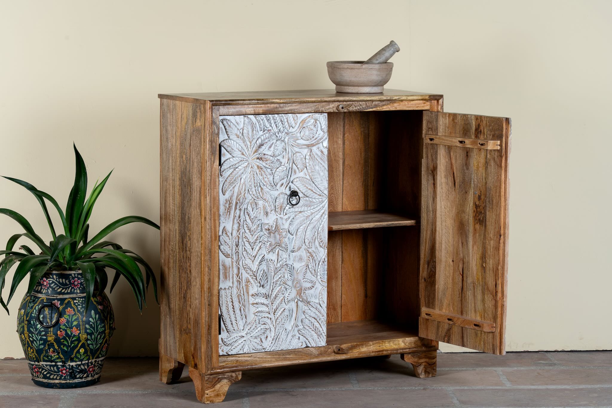 Side angle image with open door of a hand-carved mango wood cupboard Utkarsh with whitewashed doors featuring palm tree and botanical motifs. A Mediterranean-inspired piece of Indian furniture combining artisan detail with practical storage.