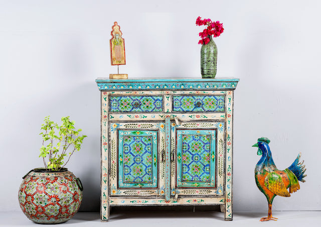 A hand-painted vintage cupboard with floral and geometric patterns in vibrant shades of blue, green, and red, on a distressed off-white base. The cupboard has two drawers and two doors.
