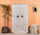 A white-washed handcrafted cabinet made from reclaimed wood, featuring intricate hand carvings and iron fittings, set against an orange wall with a potted plant to the side.