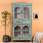 A vintage hand-carved mango wood showcase with distressed turquoise and blue finish, featuring glass panelled doors and wooden carvings, placed in a room with a plant and decorative items.