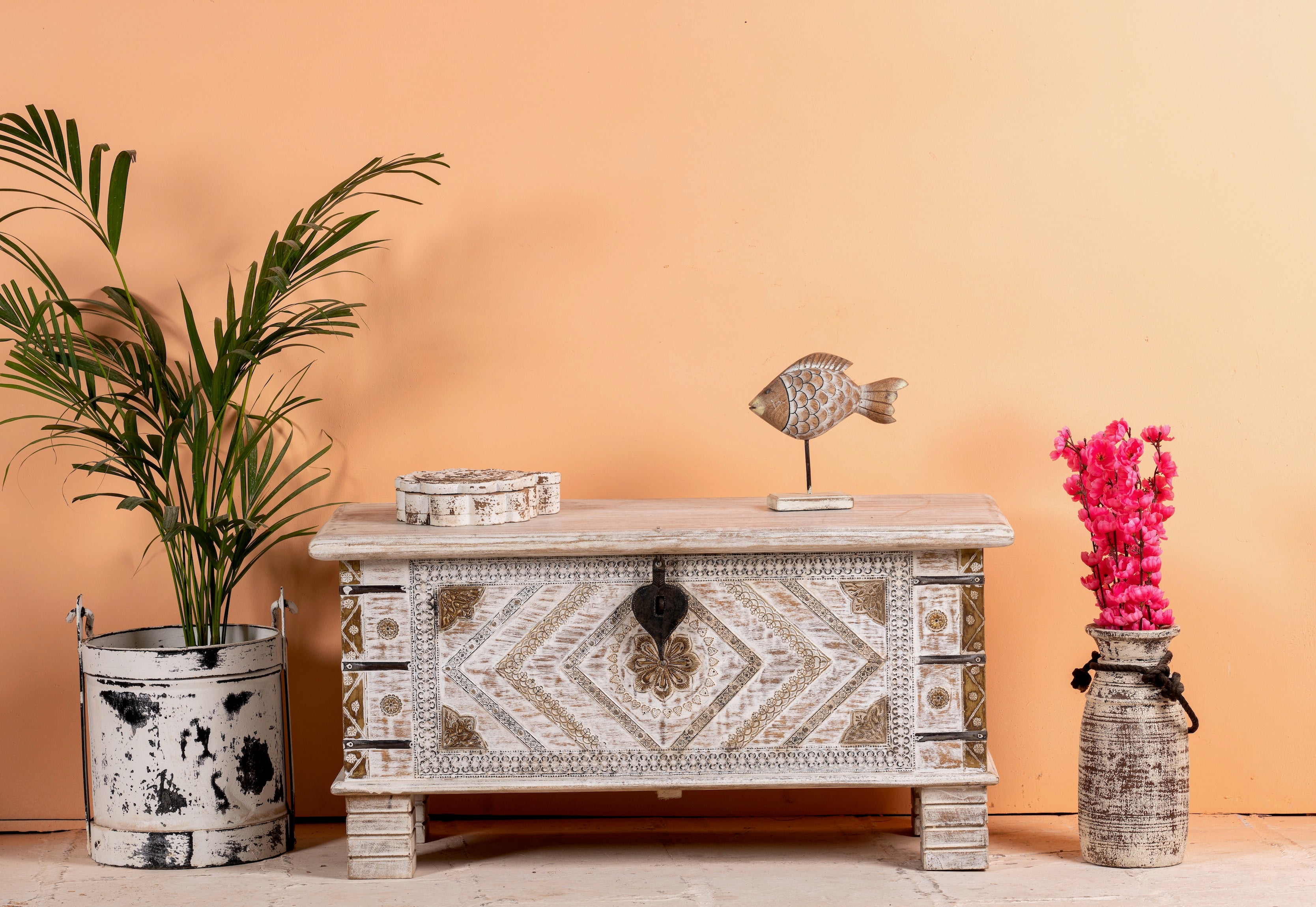A hand-carved mango wood chest with brass inlays, placed against a neutral background, flanked by a potted plant and a decorative object on its sides.